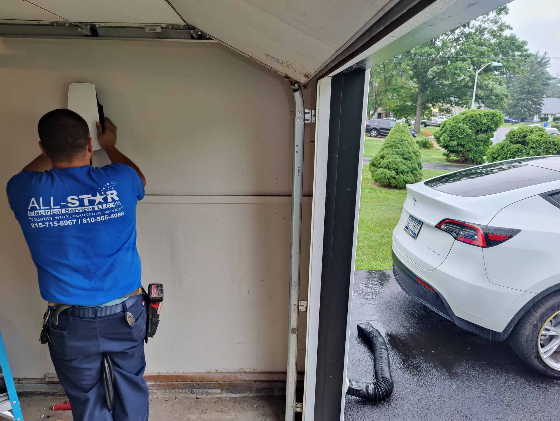 An electrician in a blue All Star Electrical Services, LLC t-shirt installing a Tesla charging station